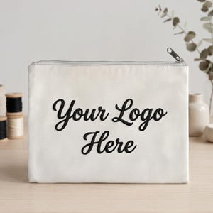 May include: Cream-coloured zippered pouch featuring the text "Your Logo Here" in black script. The rectangular pouch is on a light wooden surface. Spools of thread are visible in the background.
