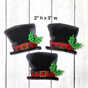 May include: Three black top hats with red and black plaid bands, gold buckles, green holly leaves, and red berries. Each hat is 2 inches high and 3 inches wide. The hats are arranged on a white wooden surface.