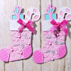 May include: Two die-cut paper Christmas stockings in shades of pink and white. Each stocking features a textured poinsettia pattern, a bright pink polka-dot toe and heel, a white cuff with a pink ribbon bow, and paper cutouts of a gift and candy cane.