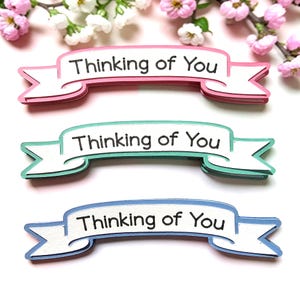 May include: Three layered banner-shaped paper cutouts in pink, teal, and blue, each with the phrase "Thinking of You" in black. The banners are white with colored edges and are arranged against a backdrop of white and pink flowers.