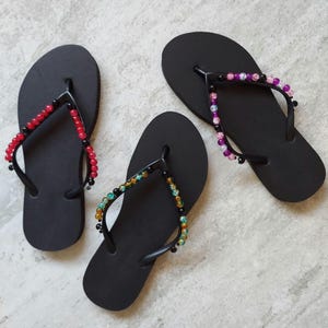 May include: Three pairs of black flip-flops with textured footbeds and Y-shaped straps. Each pair features different colored beads on the straps: one pair has red beads, one has multicolored beads, and one has purple and pink beads.