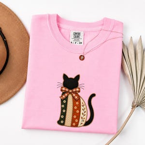 Könnte beinhalten: Ein hellrosa T-Shirt mit einem Comfort Colors-Etikett am Halsausschnitt. Das Shirt zeigt eine gestickte schwarze Katze mit einer karierten Schleife und einem Körper aus vertikalen Streifen in Grün, Creme und Orange mit Blumen- und Zickzackmustern.