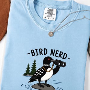 Puede incluir: Una camiseta azul claro con un gráfico de un colimbo blanco y negro de pie sobre una roca en el agua, sosteniendo unos binoculares. El texto BIRD NERD está impreso sobre el ave. La camiseta tiene una etiqueta Comfort Colors en el cuello.