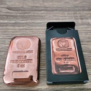 May include: A copper Germania Mint bar, 5 oz, with the text "GERMANIA MINT 999.9 FINE COPPER" and a serial number. The bar is displayed next to its black box, which also features the Germania Mint logo.