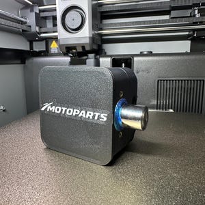 Op de afbeelding: Een zwart Motoparts-onderdeel met een zilveren mondstuk staat op het bed van een 3D-printer. Het Motoparts-logo is wit gedrukt. De printer is een Bambu Lab-model.