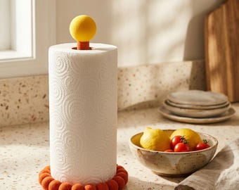 Può includere: Un portarotolo da cucina caratterizzato da un'asta verticale con una sfera giallo brillante sulla sommità e una base circolare bordata da piccole sfere in feltro color arancione bruciato. Sostiene un rotolo di carta bianca con motivi a spirale.
