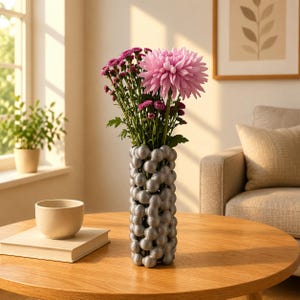 May include: A silver vase with a unique bubble design holds a bouquet of pink and purple flowers. The vase sits on a round wooden table, next to a small beige bowl and a closed book. The scene is illuminated by natural light.