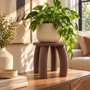 May include: A small, dark brown, three-legged stool with a smooth, rounded top. A speckled, off-white ceramic planter sits on the stool, containing a lush, green, trailing pothos plant with variegated leaves.
