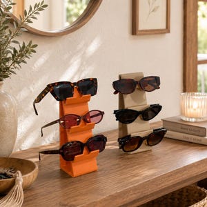 May include: Two vertical tiered display stands for eyewear, one in bright orange and one in beige, each holding three pairs of sunglasses. The orange stand features tortoiseshell, reddish-tinted, and dark frames, while the beige stand holds dark frames.