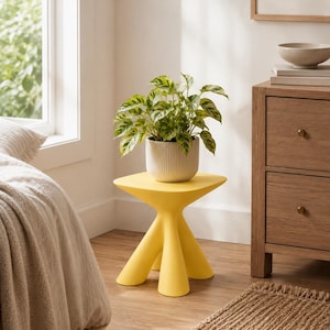 Puede incluir: Una mesa auxiliar de color amarillo brillante con una superficie cuadrada y cuatro patas gruesas y redondeadas. Sobre la mesa hay una maceta de cer&aacute;mica blanca con estr&iacute;as verticales y una planta de interior verde y blanca.