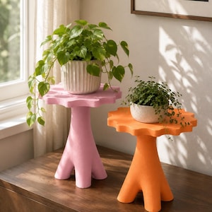May include: Two pedestal plant stands with scalloped edges, one in light pink and one in bright orange. Each stand features a central column base with three feet. A green leafy plant sits in a ribbed white pot on the pink stand, and a small green plant sits in a speckled white bowl on the orange stand.