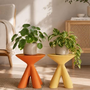 Puede incluir: Dos taburetes pequeños de tres patas con una superficie superior acampanada, uno en naranja mate y otro en amarillo mate. Cada taburete sostiene una maceta de cerámica gris claro con una planta verde, una de hojas redondas y otra variegada.