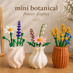 May include: Three mini botanical flower displays featuring colourful plastic brick-built flowers in textured vases. The left vase is light pink, the centre is white, and the right is terracotta. The flowers include daisies, lavender, and yellow sprigs.