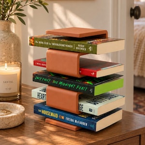May include: A terracotta-colored metal book rack featuring a modular, stacked design with three U-shaped shelves holding four books, including titles such as The Book Club for Troublesome Women, The Midnight Feast, and The Housemaid.