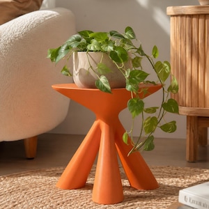 Puede incluir: Una mesa auxiliar de color naranja brillante con tres patas, acabado mate liso y tablero circular. Sobre la mesa hay una maceta de cer&aacute;mica gris claro texturizada que contiene una planta potos verde con hojas acorazonadas.