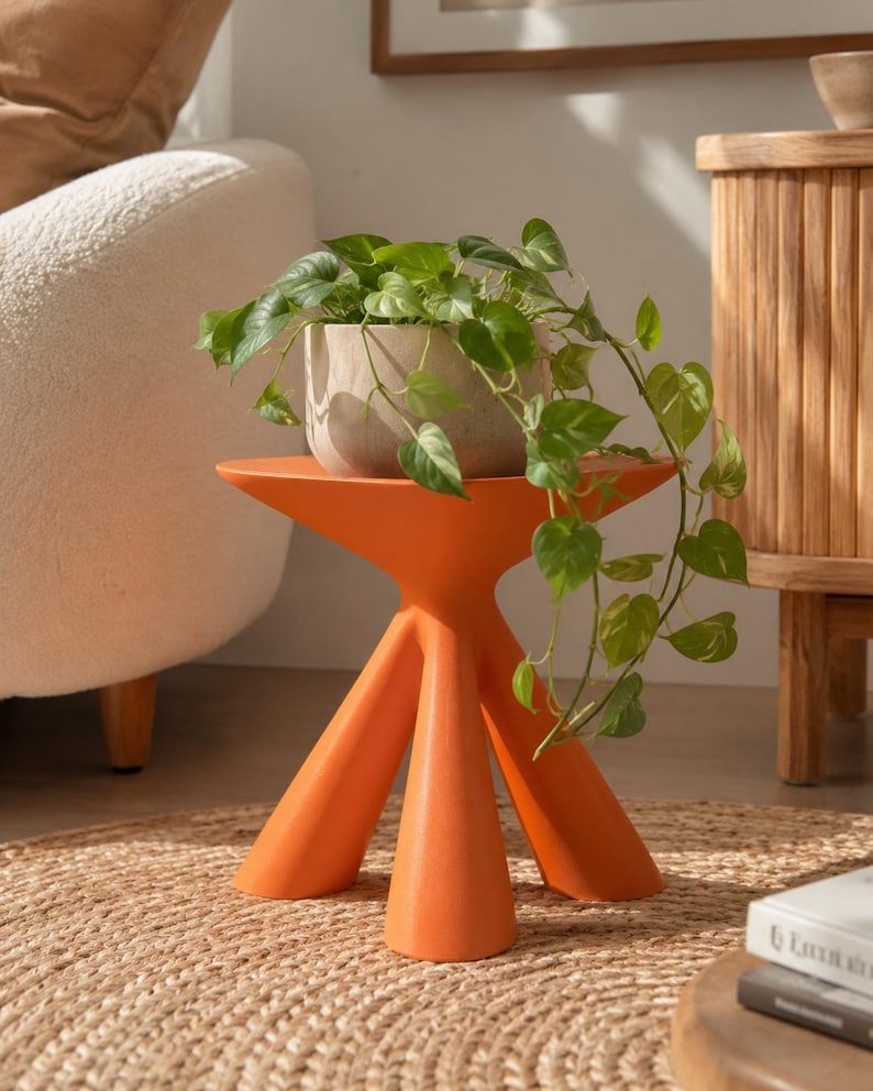 Puede incluir: Una mesa auxiliar de color naranja brillante con tres patas, acabado mate liso y tablero circular. Sobre la mesa hay una maceta de cer&aacute;mica gris claro texturizada que contiene una planta potos verde con hojas acorazonadas.