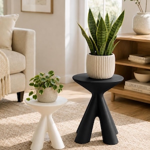 Puede incluir: Un juego de dos mesas auxiliares con dise&ntilde;o de tr&iacute;pode, una en blanco mate y otra en negro mate. Cada mesa presenta una superficie circular lisa y tres patas gruesas y c&oacute;nicas. Las mesas se muestran como un par, destacando sus colores distintos.