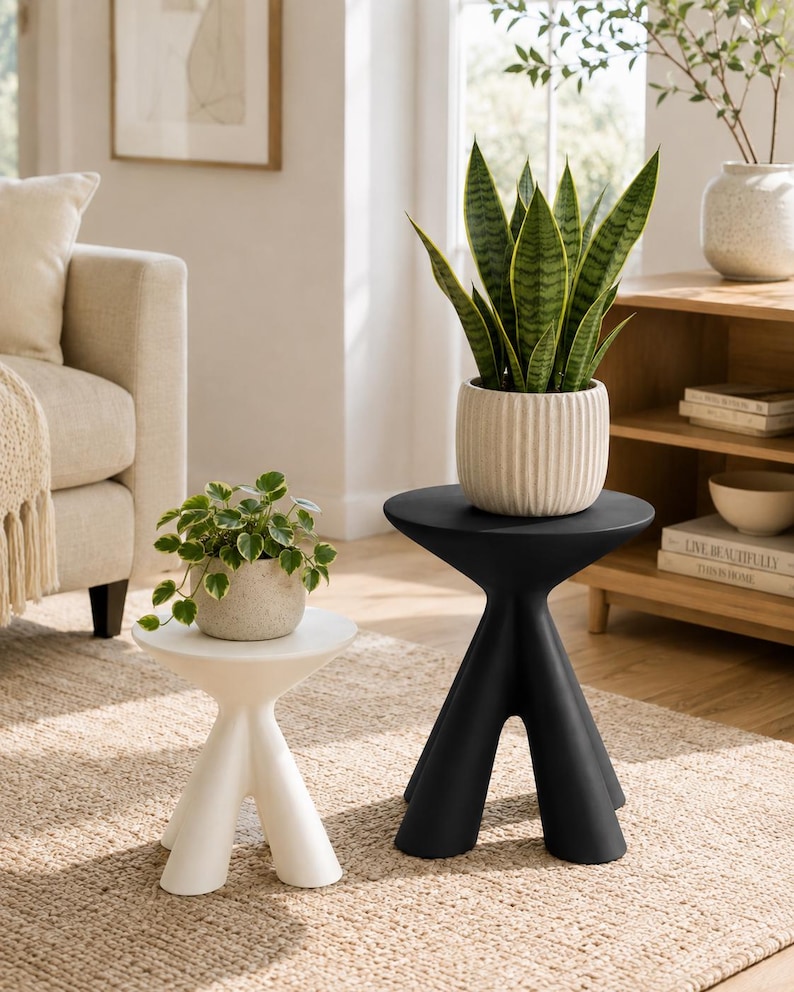 Puede incluir: Un juego de dos mesas auxiliares con dise&ntilde;o de tr&iacute;pode, una en blanco mate y otra en negro mate. Cada mesa presenta una superficie circular lisa y tres patas gruesas y c&oacute;nicas. Las mesas se muestran como un par, destacando sus colores distintos.