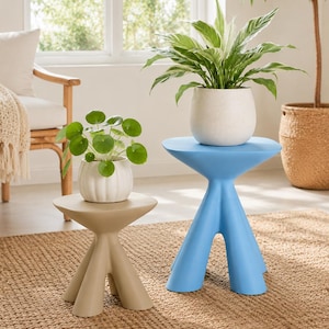 Puede incluir: Una mesa auxiliar beige de tres patas con tablero redondo y una mesa auxiliar azul de tres patas con tablero redondo. Una maceta blanca acanalada con una planta Pilea verde descansa sobre la mesa beige, y una maceta blanca moteada con una planta verde y blanca descansa sobre la mesa azul.