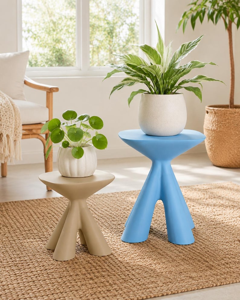 Puede incluir: Una mesa auxiliar beige de tres patas con tablero redondo y una mesa auxiliar azul de tres patas con tablero redondo. Una maceta blanca acanalada con una planta Pilea verde descansa sobre la mesa beige, y una maceta blanca moteada con una planta verde y blanca descansa sobre la mesa azul.