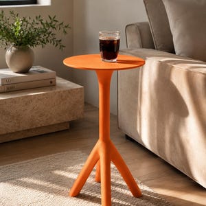 Puede incluir: Una mesa auxiliar de color naranja brillante con una superficie circular lisa y un soporte central que se divide en tres patas robustas y redondeadas. La mesa tiene un acabado mate y una estructura limpia, sosteniendo un vaso transparente con líquido oscuro.