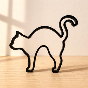 Pode incluir: Silhueta preta de um gato, feita de um material sólido, em pé sobre uma superfície de madeira. O contorno do gato é suave e contínuo, com uma cauda enrolada e orelhas pontudas. O design minimalista cria uma estética moderna.