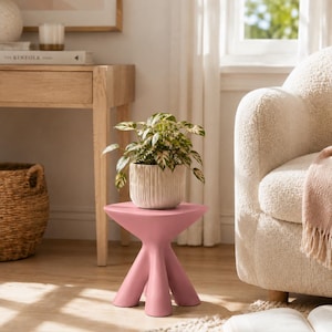 Puede incluir: Una mesa auxiliar de color rosa mate con una superficie circular plana y lisa y tres patas gruesas y redondeadas que se ensanchan hacia afuera. Una peque&ntilde;a planta verde y blanca variegada descansa en una maceta de cer&aacute;mica estriada.