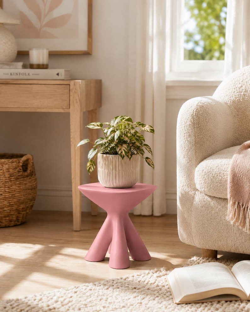 Puede incluir: Una mesa auxiliar de color rosa mate con una superficie circular plana y lisa y tres patas gruesas y redondeadas que se ensanchan hacia afuera. Una peque&ntilde;a planta verde y blanca variegada descansa en una maceta de cer&aacute;mica estriada.