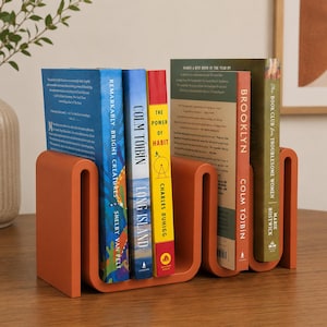 Puede incluir: Un sujetalibros de metal naranja mate en forma de S que sostiene cinco libros en posición vertical. Los libros incluyen títulos como Remarkably Bright Creatures, Long Island, The Power of Habit, Brooklyn y The Book Club for Troublesome Women, alineados.