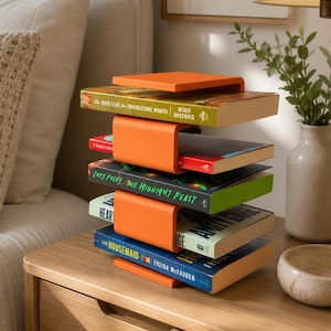 Puede incluir: Un estante para libros vertical de metal naranja con una serie de repisas en forma de U que sostienen cuatro libros apilados horizontalmente. Los libros incluyen títulos como The Book Club for Troublesome Women y The Housemaid.