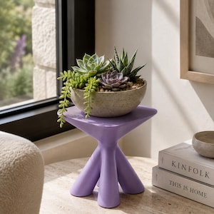Puede incluir: Un taburete de color morado claro con tres patas y una parte superior triangular sostiene un cuenco moteado de color claro lleno de una variedad de suculentas verdes y moradas, incluida una planta colgante, dispuestas sobre guijarros.