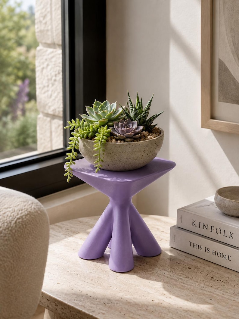 Puede incluir: Un taburete de color morado claro con tres patas y una parte superior triangular sostiene un cuenco moteado de color claro lleno de una variedad de suculentas verdes y moradas, incluida una planta colgante, dispuestas sobre guijarros.
