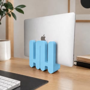 May include: A light blue, wavy-shaped vertical laptop stand holding a silver laptop in an upright position. The stand features a continuous, rounded, serpentine design that supports the device securely while it rests on a flat surface.