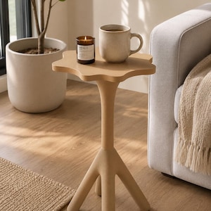 May include: A beige side table with a flower-shaped top and three thick, tapered legs. On the table sits a lit candle in a dark glass jar with a white label reading SANTAL 26 and a speckled, light-colored ceramic mug with a handle.