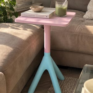 May include: A square side table with a light pink top and a matching pink central column that transitions into a light blue three-legged base. The table surface holds a white book titled At Home in the World, a small stone bowl, and a ribbed glass.