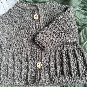 May include: A grey crochet baby cardigan with two beige buttons. The handmade sweater features a textured stitch pattern and a round neckline. The sleeves are long and the bottom hem has a slight flare. The cardigan is displayed on a green fabric background.