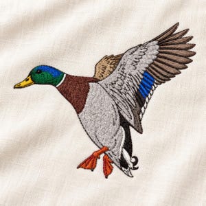 May include: An embroidered mallard duck in flight on a light beige fabric. The duck features a vibrant green head, a yellow beak, a brown chest, and wings with grey, white, and blue feathers. The duck has bright orange webbed feet.