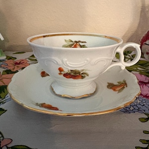 May include: White teacup and saucer set with gold trim and fruit illustrations. The teacup has a handle and sits on a matching saucer. The set is likely porcelain and is a vintage or antique item. The fruit illustrations are in shades of orange and green.