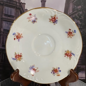May include: White porcelain saucer with a gold rim and floral pattern. The saucer features small clusters of red, blue, and yellow flowers. The saucer is displayed on a wooden stand, showcasing its elegant design. A classic piece for serving tea or desserts.