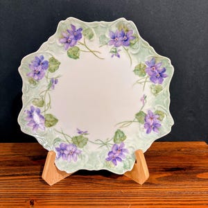 May include: A white porcelain plate with a scalloped edge featuring a hand-painted pattern of purple violet flowers and green leaves. The floral design is arranged in a circular border around the center of the plate, which has a light green wash.