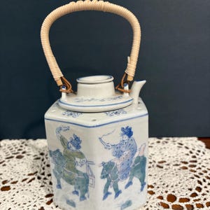 May include: A hexagonal ceramic teapot featuring blue and green hand-painted illustrations of figures riding horses. The teapot has a white glazed finish, a matching lid, and a curved handle wrapped in light tan woven material for a secure grip.