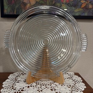 May include: A clear glass serving platter featuring a concentric circular ribbed pattern across its surface. The round platter includes two integrated side handles with vertical ridges, providing a textured grip on both the left and right sides.