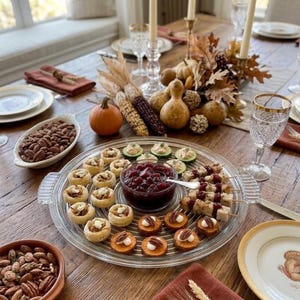May include: A clear glass serving platter holds an assortment of appetizers including small pastry cups, cucumber slices topped with spread, skewered meat bites with cranberry sauce, and sweet potato rounds with pecans.