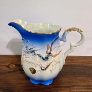 May include: A porcelain pitcher featuring a gradient blue and cream finish with a raised, textured dragon motif in shades of white, black, and gold. The vessel has a scalloped rim, a curved handle with gold trim, and a glossy, iridescent interior.