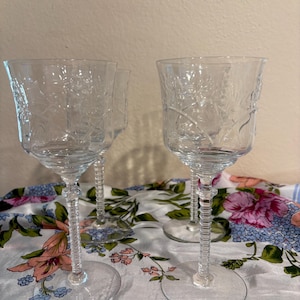 May include: Four clear, stemmed wine glasses with etched floral designs. The glasses have a round base, a twisted stem, and a flared bowl. They are arranged on a floral patterned tablecloth.