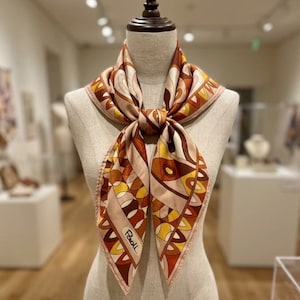 May include: A silk scarf with a geometric pattern in shades of brown, yellow, and beige, knotted at the neck. The scarf has a pink border and the name "Paoll" is printed on the bottom corner. The scarf is displayed on a mannequin.