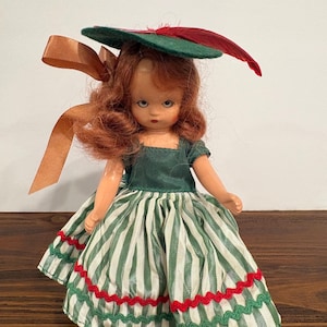 May include: A doll with reddish-brown hair wearing a green hat with a red feather and a brown ribbon. The doll has a dark green bodice and a skirt featuring vertical white and green stripes, accented with horizontal red and green wavy trim.