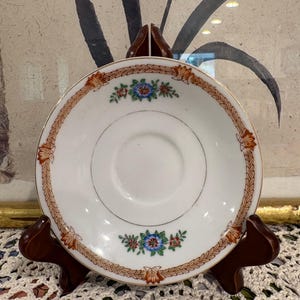May include: A white saucer with a gold rim and a decorative border of brown leaves and a floral design. The saucer is supported by a wooden stand, and the background features a painting.