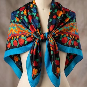 May include: A colorful square scarf with a floral pattern. The scarf features a black background with red, orange, and blue flowers. It has a bright blue border and is tied in a knot. The scarf is displayed on a mannequin.