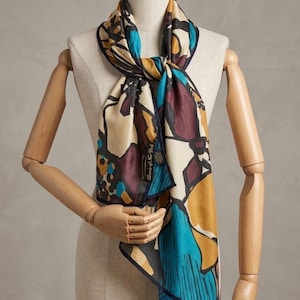 May include: A silk scarf with a colorful floral pattern in shades of beige, teal, burgundy, and gold. The scarf is draped on a wooden mannequin. The scarf has a dark blue border and a label that reads "Burns of New York".